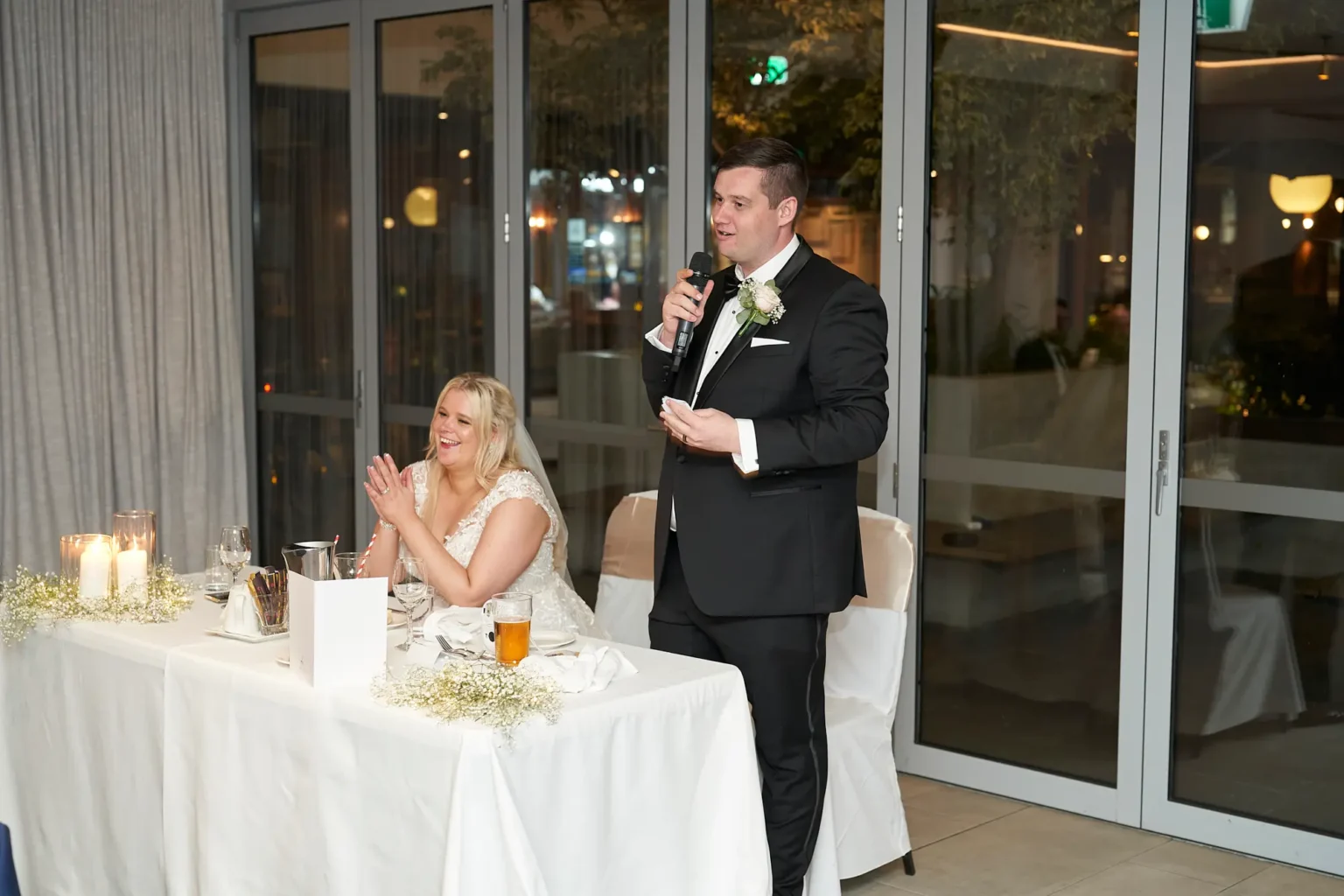 Groom delivers a heartfelt speech during the wedding reception at Amora Herencia Riverwalk Melbourne while the bride sits beside him smiling and clapping with pride. Standing at their sweetheart table decorated with baby’s breath flowers and candles, the groom in a black tuxedo speaks to guests, capturing an emotional and joyful moment of the bride and groom’s Melbourne wedding celebration.
