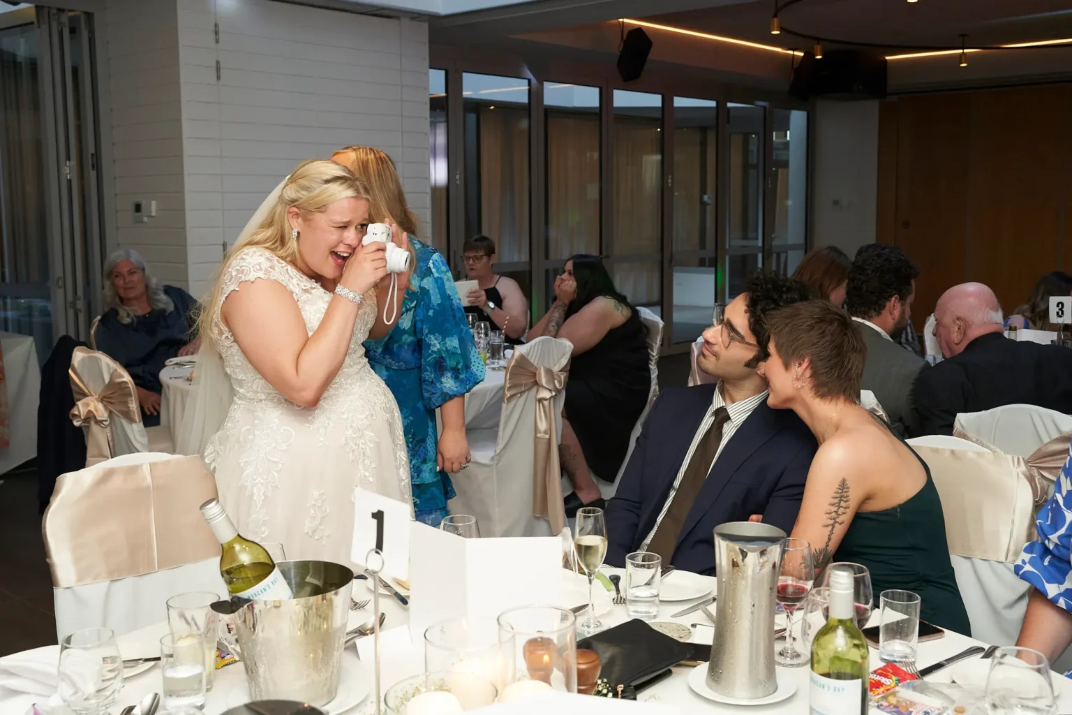 Bride laughs while taking a fun instant photo of guests with a Fujifilm Instax camera during the wedding reception at Amora Herencia Riverwalk Melbourne. Leaning toward a guest table decorated with candles, wine glasses, and white linens, the bride in her lace wedding gown creates a playful and candid moment celebrating with friends and family.