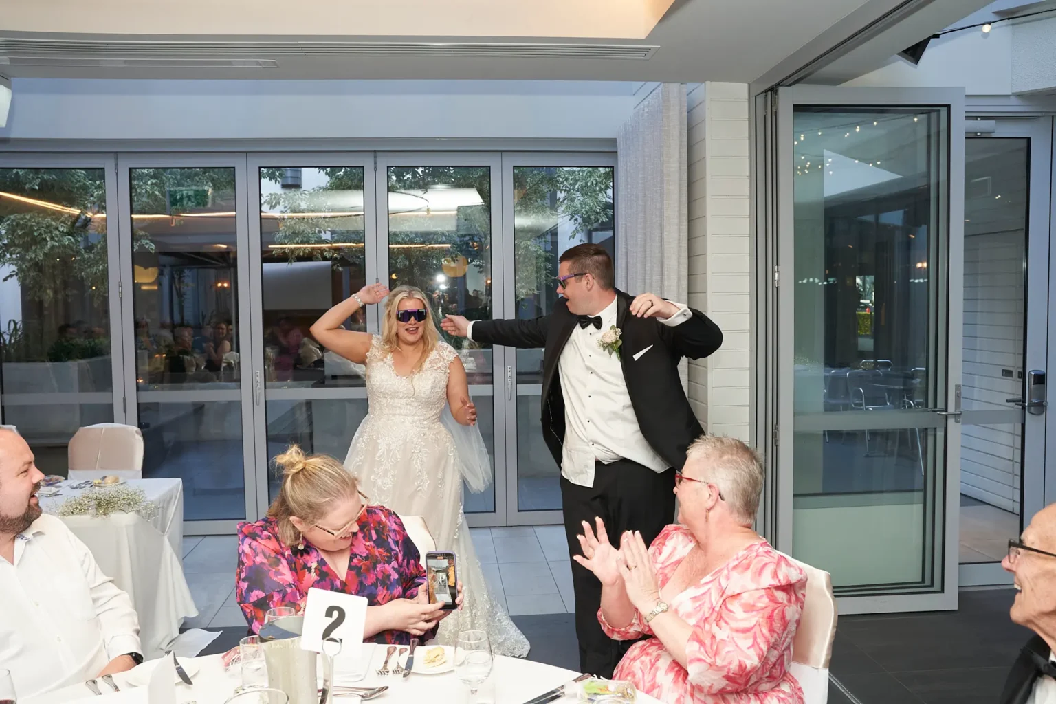 Bride and groom enter Amora Herencia Riverwalk Melbourne wedding reception wearing blue party sunglasses as guests cheer and clap, capturing a joyful grand entrance in the bright Event Pavilion dining room.