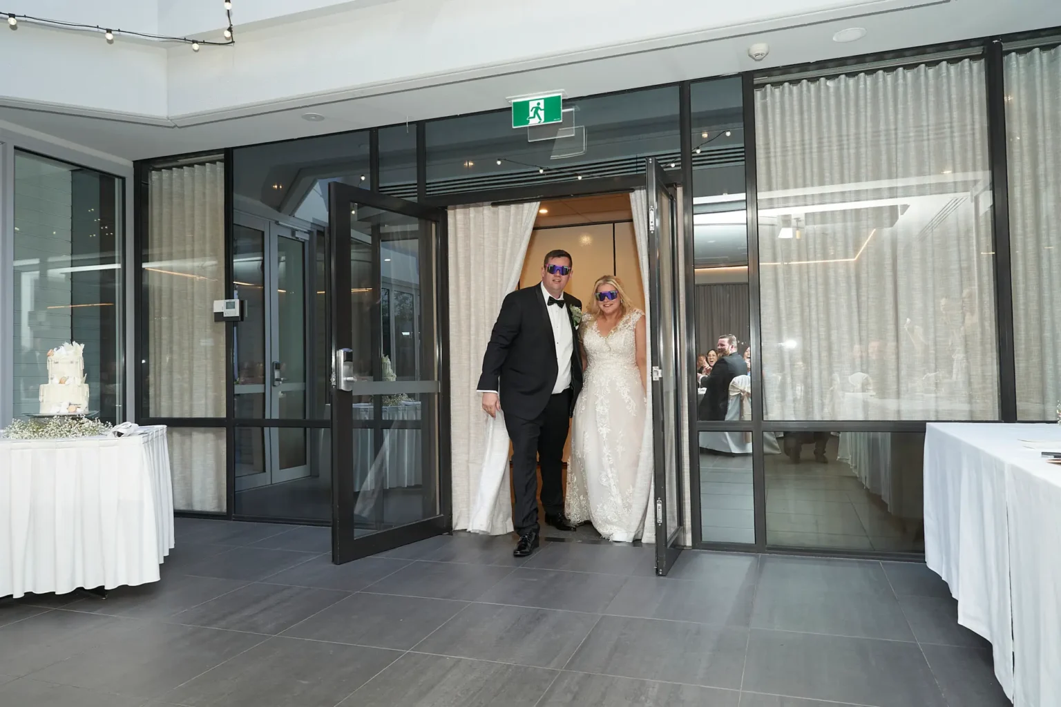 Bride and groom make a joyful grand entrance at Amora Herencia Riverwalk Melbourne wedding reception wearing blue party sunglasses, walking hand in hand through glass doors as guests cheer inside the bright Event Pavilion reception space.