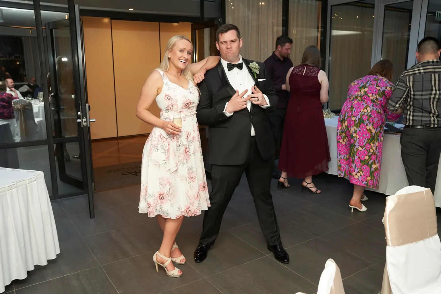Groom shares a playful moment with a guest during the wedding reception at Amora Herencia Riverwalk Melbourne, posing with a fun hand gesture while the guest laughs with a glass of champagne. Surrounded by other guests and reception décor, the candid moment captures the lively and joyful party atmosphere of the Melbourne wedding celebration.