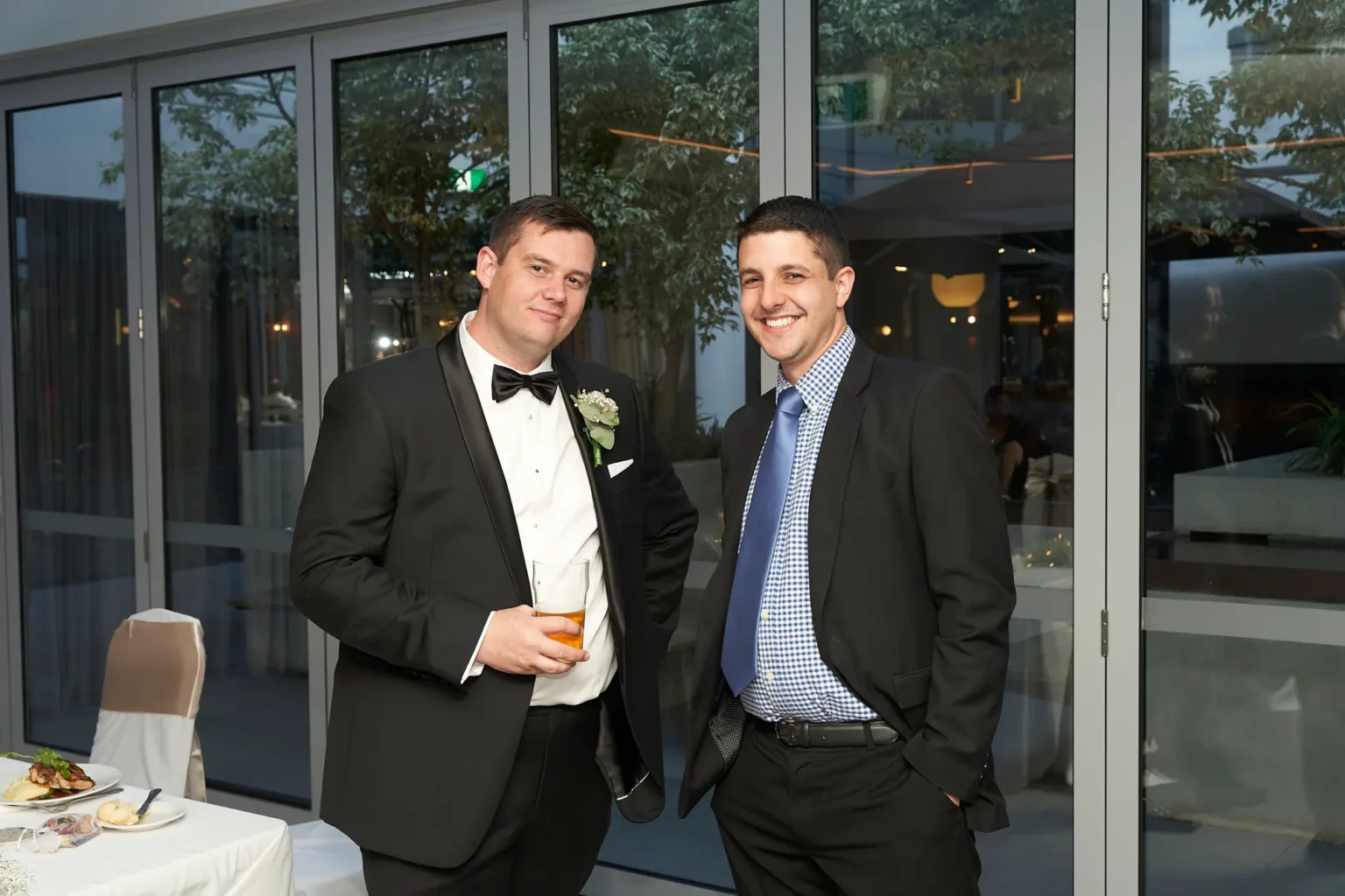 Groom stands beside a smiling guest during the wedding reception at Amora Herencia Riverwalk Melbourne, both holding beers and posing casually for the camera. The groom in a black tuxedo with a white rose boutonniere and his guest in a dark suit share a relaxed and friendly moment, capturing the joyful and social atmosphere of the bride and groom’s Melbourne wedding celebration.