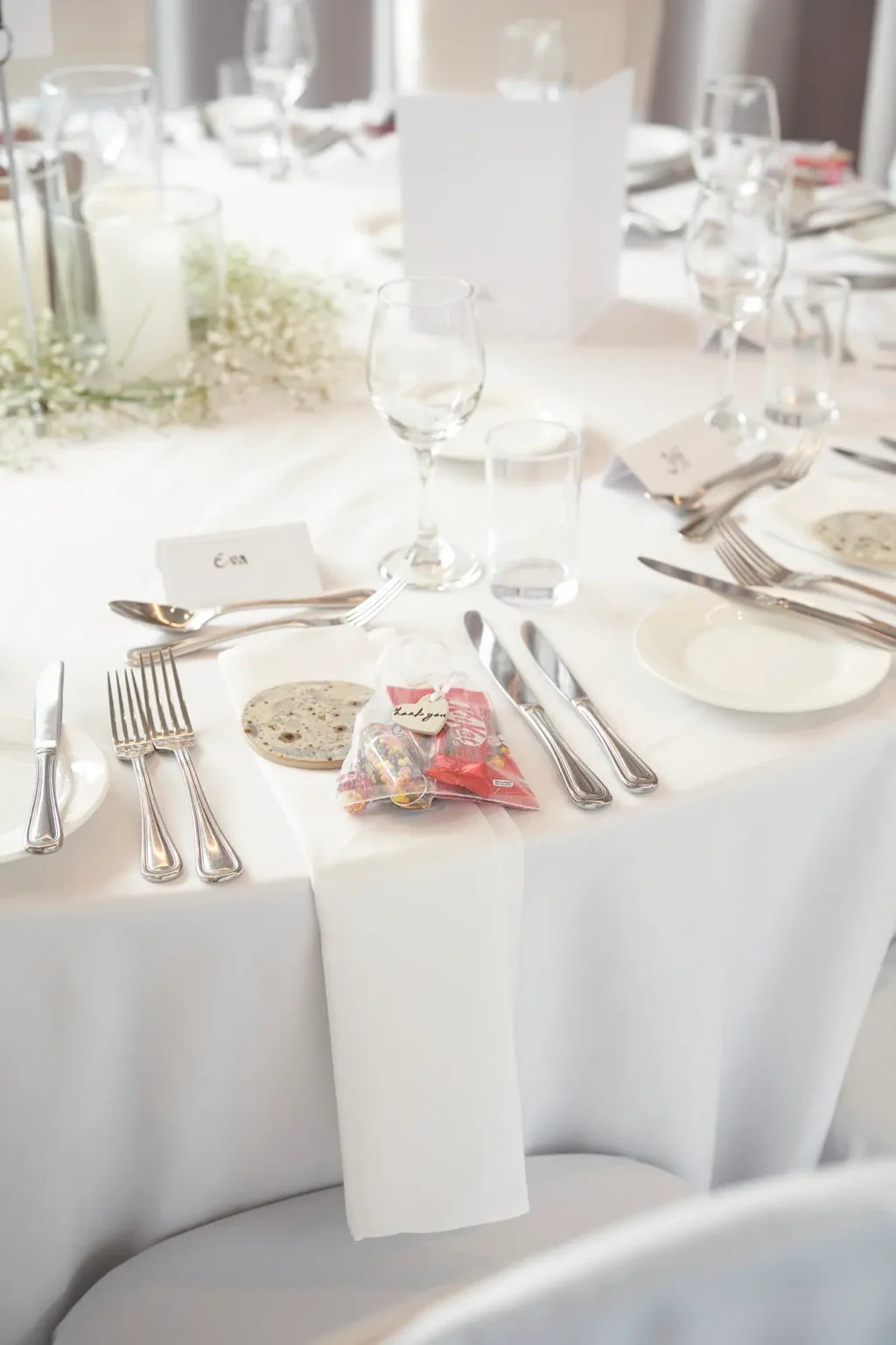 Close-up wedding reception place setting at Amora Herencia Riverwalk Melbourne featuring guest name card, polished silver cutlery, wine glass, colorful lolly favor bag with thank you tag, and baby’s breath centerpiece with candles in a bright reception room prepared for the bride and groom.