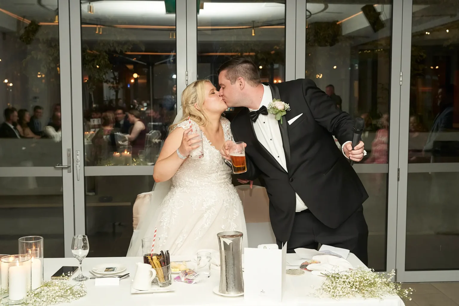 Bride and groom share a joyful kiss across their sweetheart table after the groom’s speech during the wedding reception at Amora Herencia Riverwalk Melbourne. The groom holds a microphone and beer while the bride holds a glass with a striped straw, surrounded by baby’s breath flowers and candles, capturing a celebratory moment of their Melbourne wedding reception.