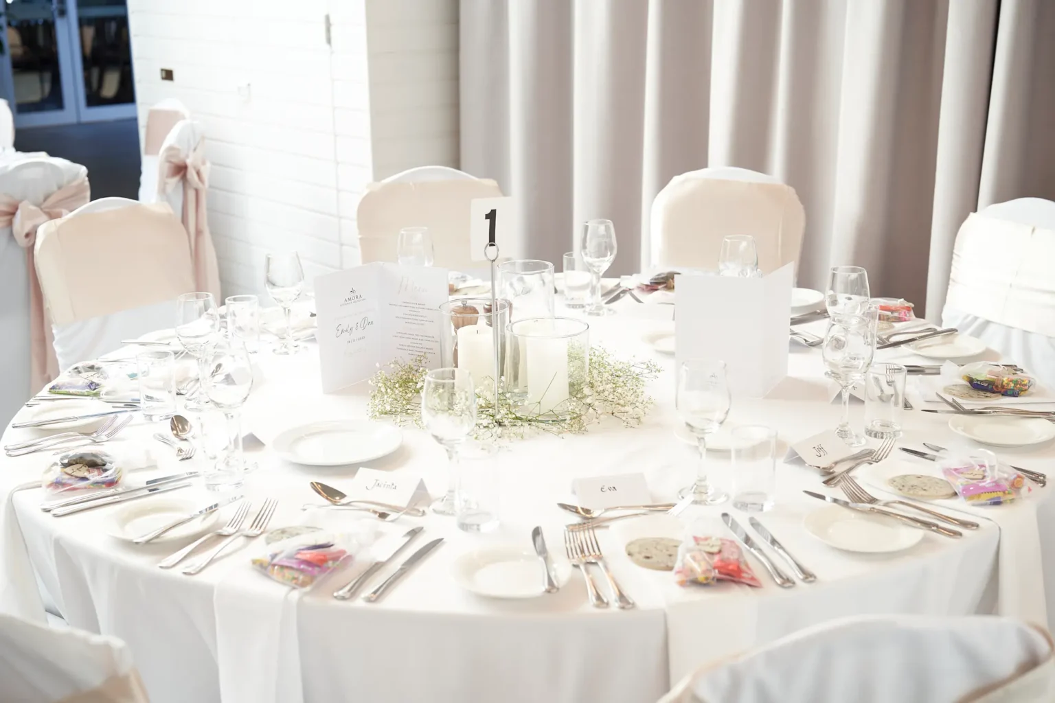 Wedding reception table setting at Amora Herencia Riverwalk Melbourne featuring baby’s breath floral centerpiece with pillar candles, white linen tablecloth, guest name cards, wedding favors, menu stationery, and champagne-gold chair bows in a bright reception room prepared for the bride and groom.