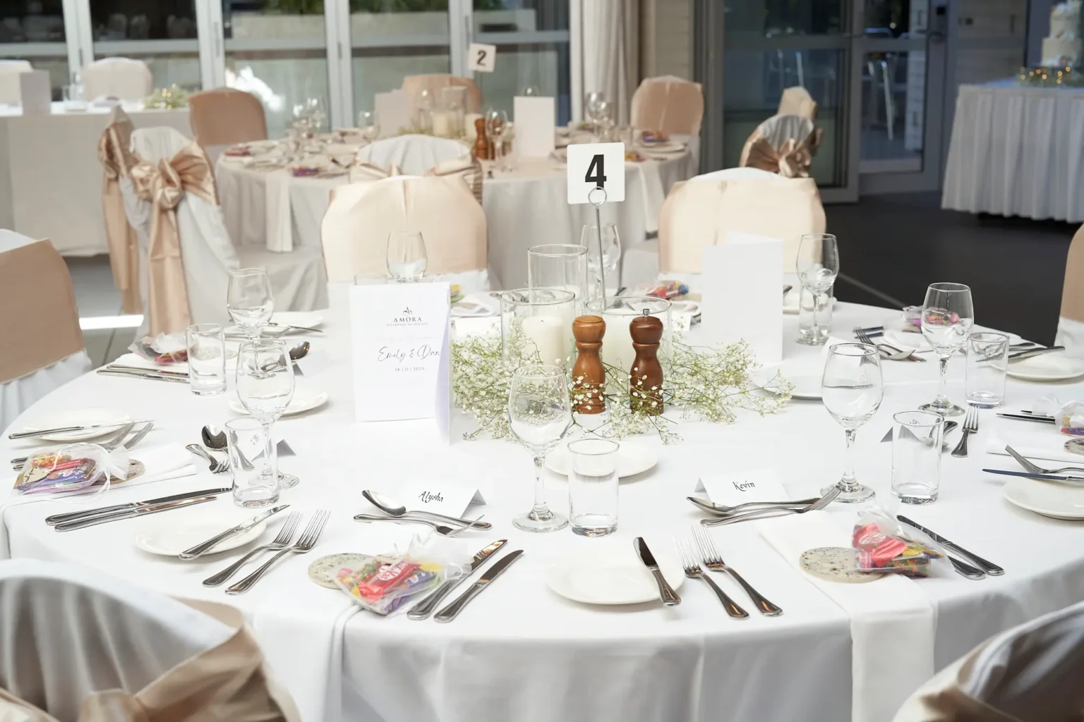 Elegant wedding reception table setup at Amora Herencia Riverwalk Melbourne featuring white linen tablecloths, baby’s breath floral centerpieces with pillar candles, personalized place cards, and guest favors. Chairs are decorated with champagne-gold bows, with natural light from large glass doors overlooking the Yarra River parklands.