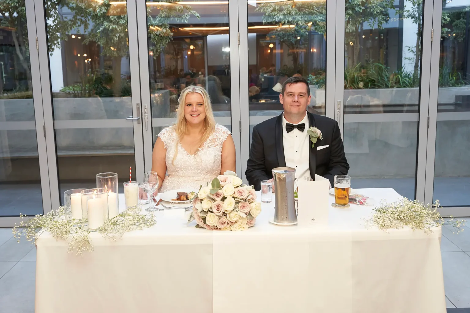 Bride and groom sit together at their sweetheart table during the wedding reception at Amora Herencia Riverwalk Melbourne, smiling toward the camera while sharing their first meal as newlyweds. The table is decorated with baby’s breath flowers, pillar candles, and a bouquet of blush and white roses, creating an intimate and elegant moment inside the bright Event Pavilion.