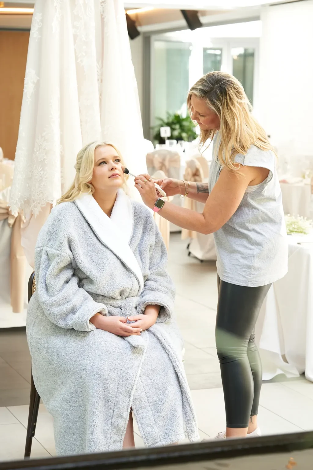 Bride sits calmly as her makeup artist applies the final touches during bridal preparations at Amora Herencia Riverwalk Melbourne. Wearing a soft grey fleecy robe with white trim, the bride looks slightly upward with a serene expression while the artist carefully applies makeup with a brush. Her white lace wedding gown hangs elegantly behind her, symbolising the moment before she becomes a bride. In the background, reception tables are beautifully arranged with white linens and champagne bows, capturing the quiet anticipation of a Melbourne riverside wedding morning.