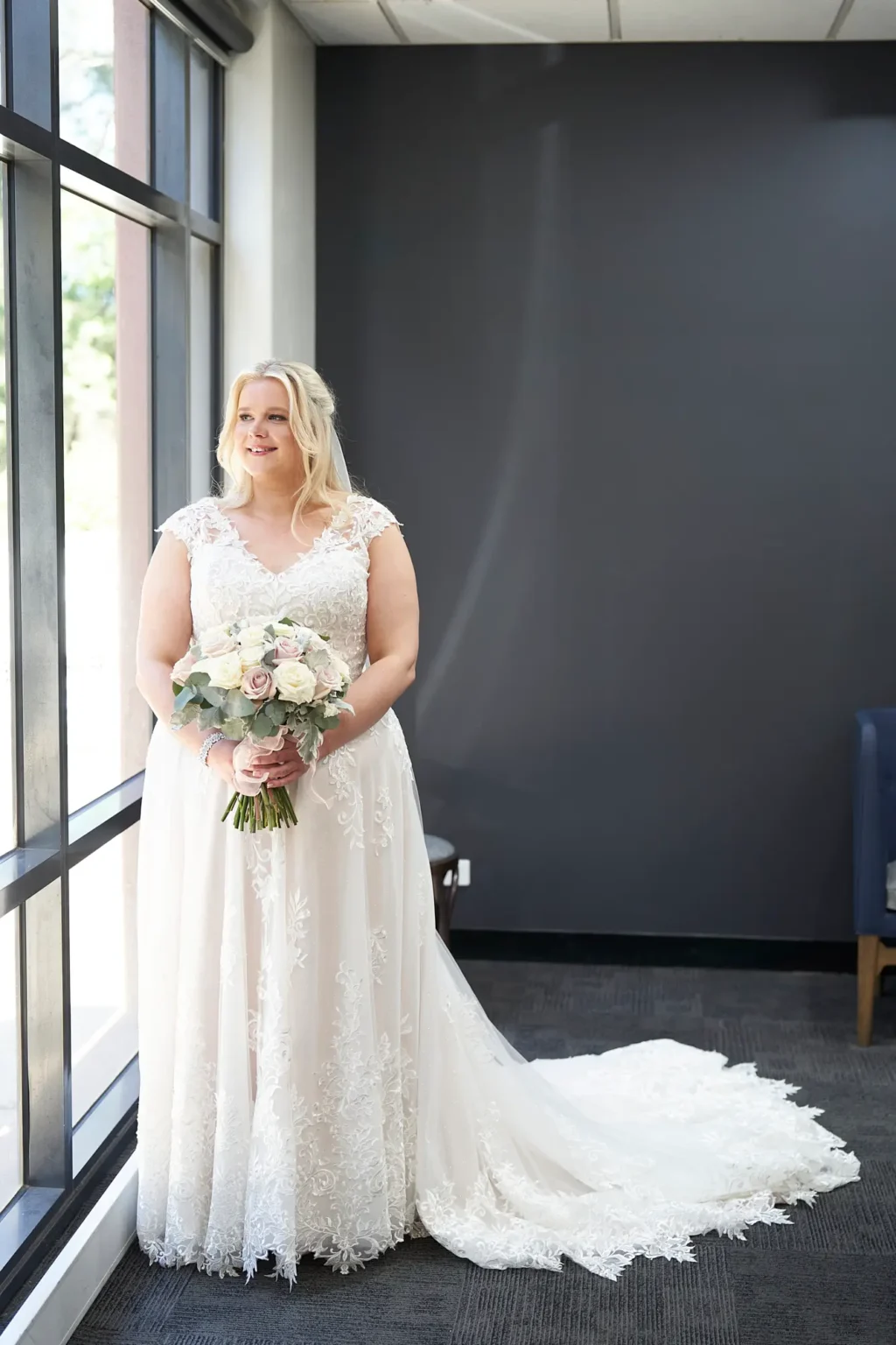 Bride stands gracefully beside a large window during bridal preparations at Amora Herencia Riverwalk Melbourne, softly looking toward the natural light with a gentle smile. She wears a cap-sleeved lace A-line wedding gown with a beautifully detailed scalloped train flowing across the floor, while holding a bouquet of blush and white roses with eucalyptus and dusty miller. The modern interior with a dark feature wall and floor-to-ceiling windows creates a striking contrast, highlighting this elegant and serene bridal portrait before the Melbourne riverside wedding ceremony.