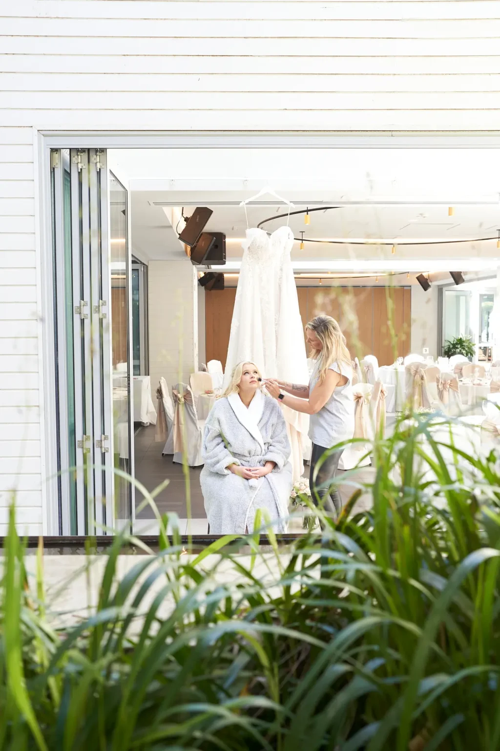 Bride sits calmly while her makeup artist applies the finishing touches during bridal preparations at Amora Herencia Riverwalk Melbourne. Wearing a soft grey robe, the bride looks upward with a serene expression as natural daylight fills the room. Her wedding gown hangs elegantly behind her, while reception tables dressed in white linens and champagne bows are visible in the background. Framed through lush garden grasses outside the window, this quiet moment captures the anticipation and intimate atmosphere of a Melbourne riverside wedding morning.