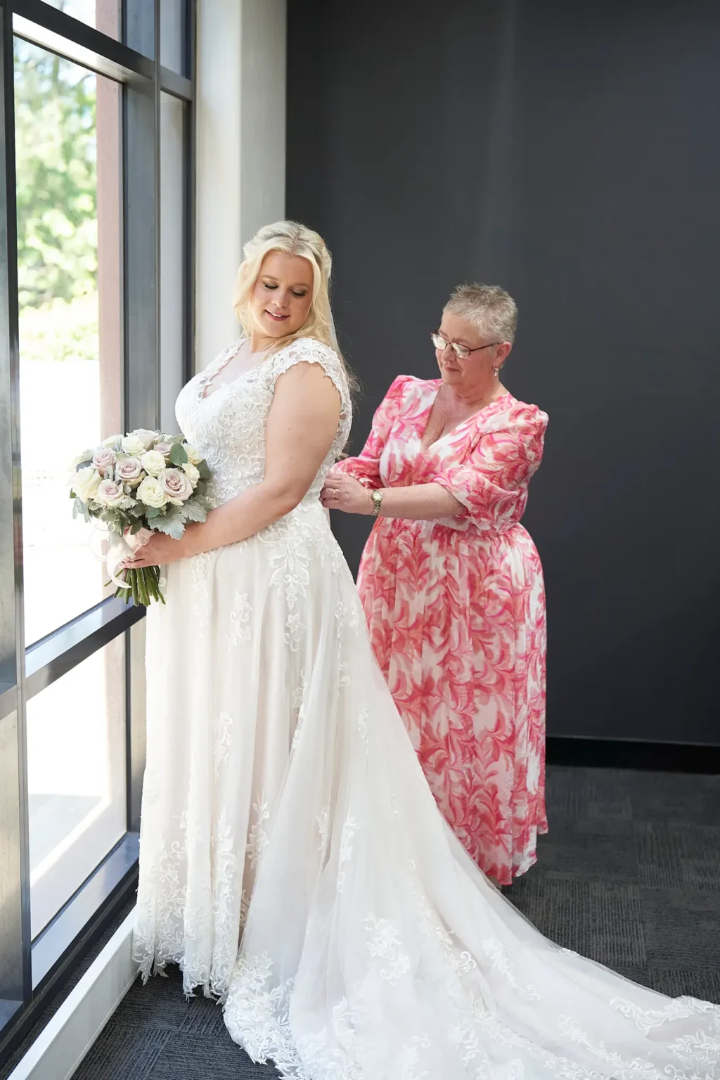 Bride stands gracefully by a sunlit window as a close family member carefully fastens the back of her lace wedding gown during bridal preparations at Amora Herencia Riverwalk Melbourne. Holding her bouquet and smiling softly, the bride is captured in profile while natural light highlights the intricate lace details and flowing train of the dress. The modern corridor setting and glimpses of green parkland outside create a calm and intimate moment, capturing the emotional final touches before the Melbourne riverside wedding ceremony.