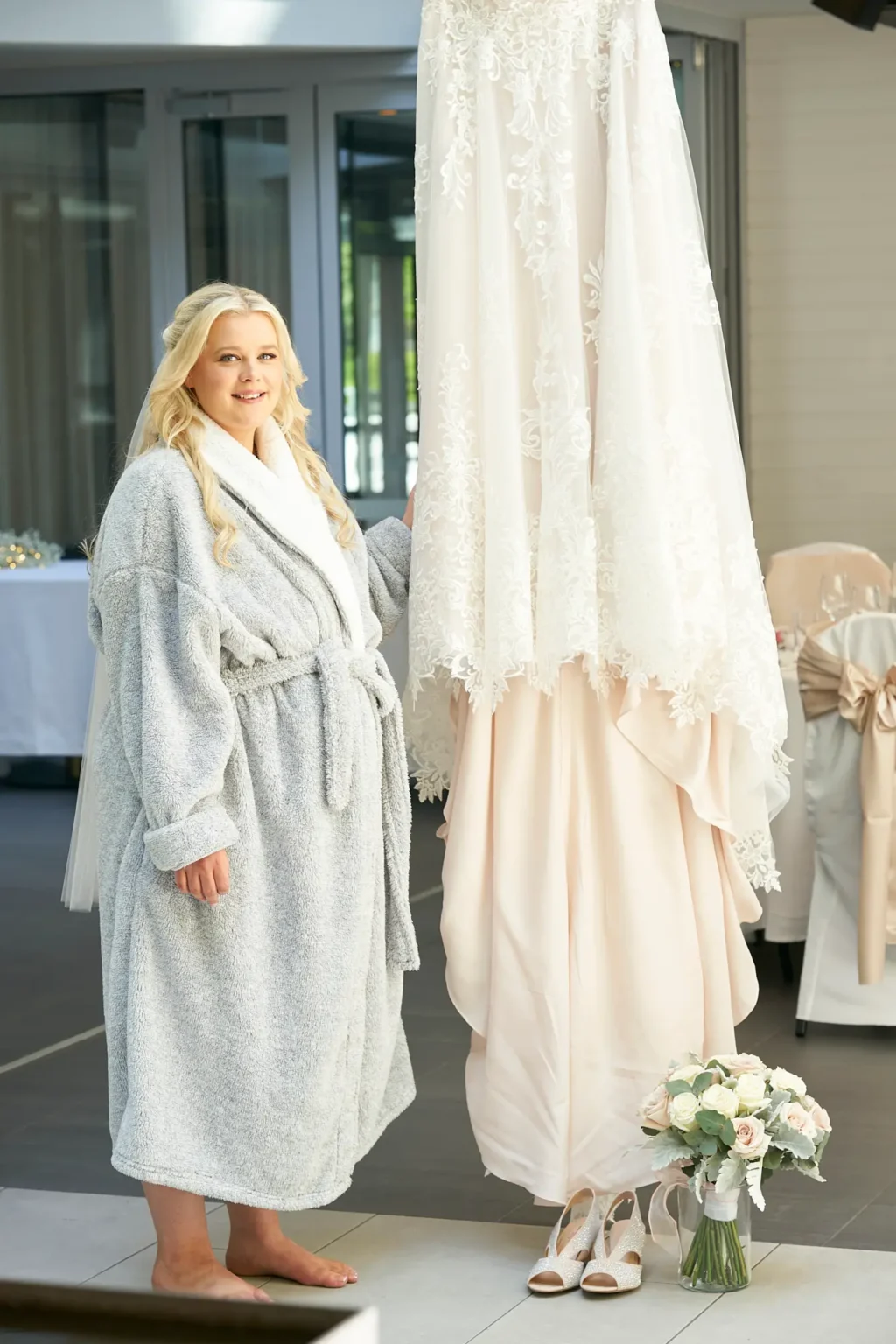 Bride stands barefoot beside her lace wedding gown during a quiet moment of bridal preparations at Amora Herencia Riverwalk Melbourne. Wearing a soft grey robe with her hair styled in elegant waves and a delicate veil, she gently rests her hand on the gown while smiling calmly toward the camera. Her bridal shoes and bouquet of blush and white roses sit beneath the dress, while the reception room behind is set with white linen tables and champagne ribbon chairs, capturing the peaceful anticipation before the Melbourne riverside wedding ceremony.