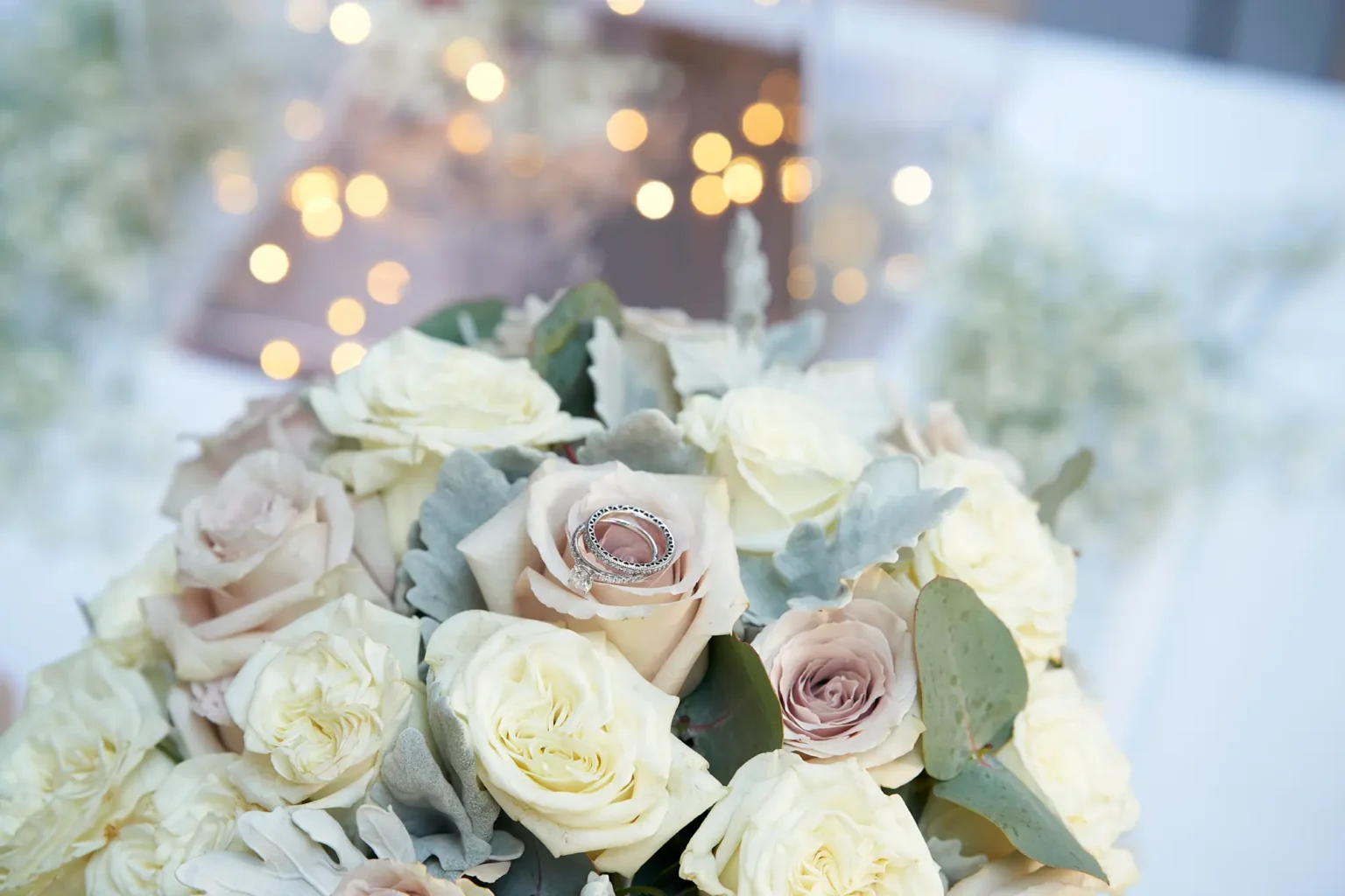 Macro wedding detail of the bride and groom’s diamond engagement ring and wedding bands delicately balanced on a blush rose within the bridal bouquet at Amora Herencia Riverwalk Melbourne. Surrounded by soft cream and dusty pink roses, eucalyptus, and dusty miller leaves, the rings sparkle as warm golden fairy lights blur into a dreamy bokeh background. The shallow depth of field highlights the textures of the petals and jewellery, capturing an elegant and romantic detail from this intimate Melbourne riverside wedding.
