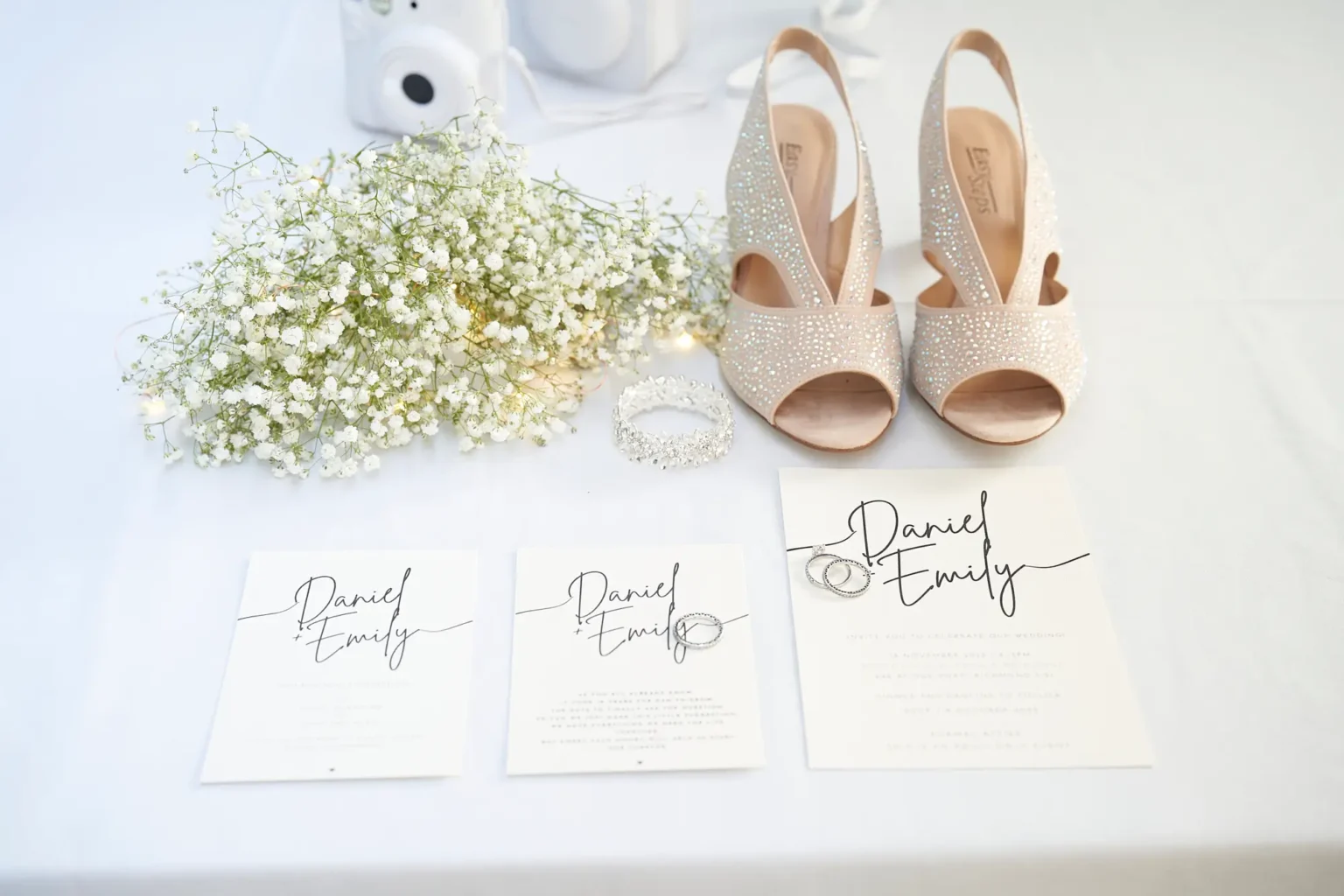Elegant wedding flat lay featuring the bride and groom’s invitation suite with their wedding rings resting on the card, photographed during wedding preparations at Amora Herencia Riverwalk Melbourne. Crystal peep-toe bridal heels, a sparkling bracelet, and a white Fujifilm Instax Mini camera are carefully arranged on soft white linen. Baby’s breath flowers and delicate fairy lights add a romantic touch, capturing the thoughtful details and anticipation of a Melbourne riverside wedding day before the ceremony begins.