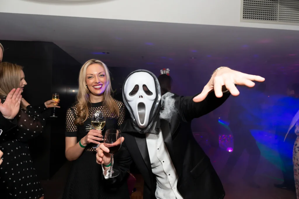 svg%3E A playful moment at the Kooyong Tennis Club Halloween Gala in Melbourne shows a man in a black tuxedo wearing a Ghostface mask lunging toward the camera, a wisp of smoke adding a ghostly effect, while a woman in a black cocktail dress laughs and holds two glasses of wine, capturing the elegant yet spooky atmosphere of this luxury end-of-year ball with colorful ambient lighting and joyful guest interactions.