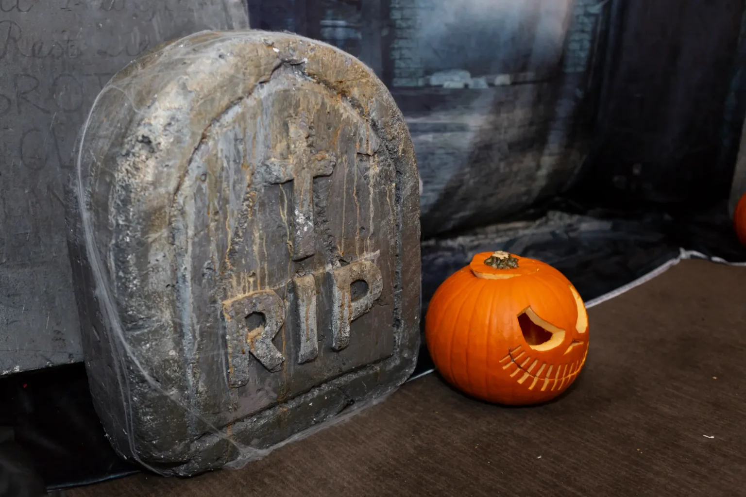 A styled graveyard photo backdrop at the Kooyong Tennis Club Halloween Gala features a weathered tombstone with RIP engraving draped in spiderwebs beside a carved jack-o’-lantern with a sharp-toothed grin, set against a misty cemetery scene, creating a theatrical and polished photo-op that captures the spooky yet luxurious atmosphere of this Melbourne end-of-year ball.