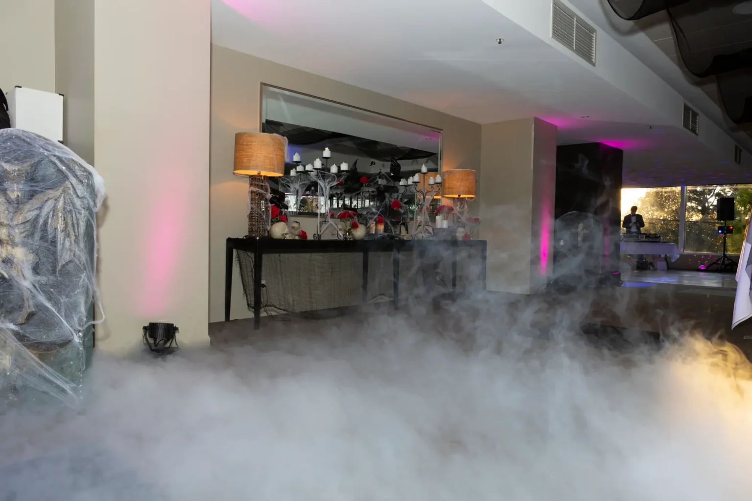 A moody indoor graveyard setup at the Kooyong Tennis Club Halloween Gala features a console table styled with candelabras, skulls, vintage lamps and deep red roses draped in thick spiderwebs, while low-lying fog blankets the floor and magenta lighting glows against a pillar, with sunset light and a DJ preparing in the background, capturing the immersive, eerie atmosphere of this luxury haunted mansion themed event in Melbourne.