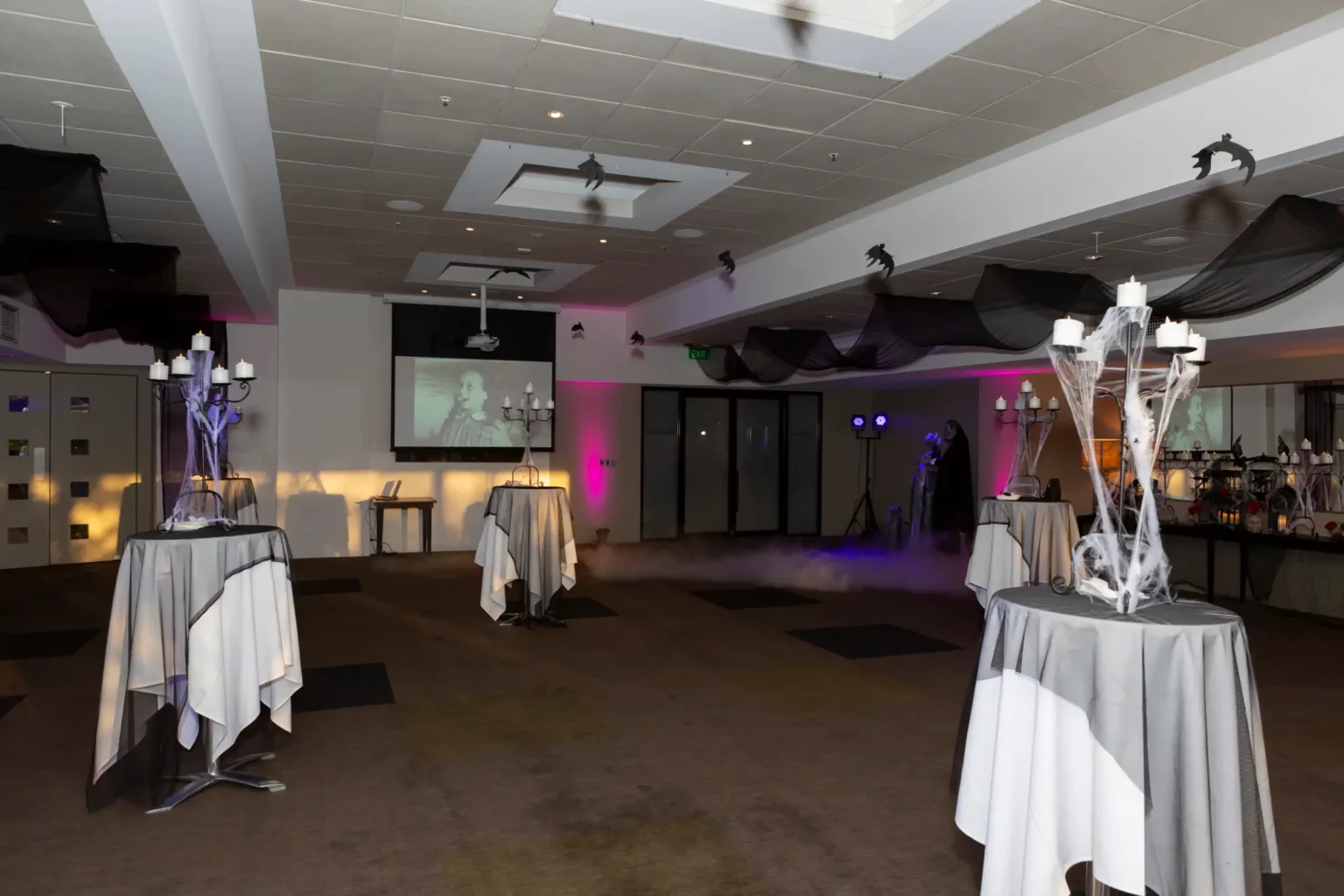 A spacious ballroom at the Kooyong Tennis Club Halloween Gala is styled with cocktail tables featuring candelabra centrepieces draped in spiderwebs, beneath black ceiling drapes and bat decorations, while low-lying fog spreads across the floor and a projection screen displays vintage horror visuals, capturing the moody, theatrical and luxurious haunted mansion atmosphere before guests arrive in Melbourne.