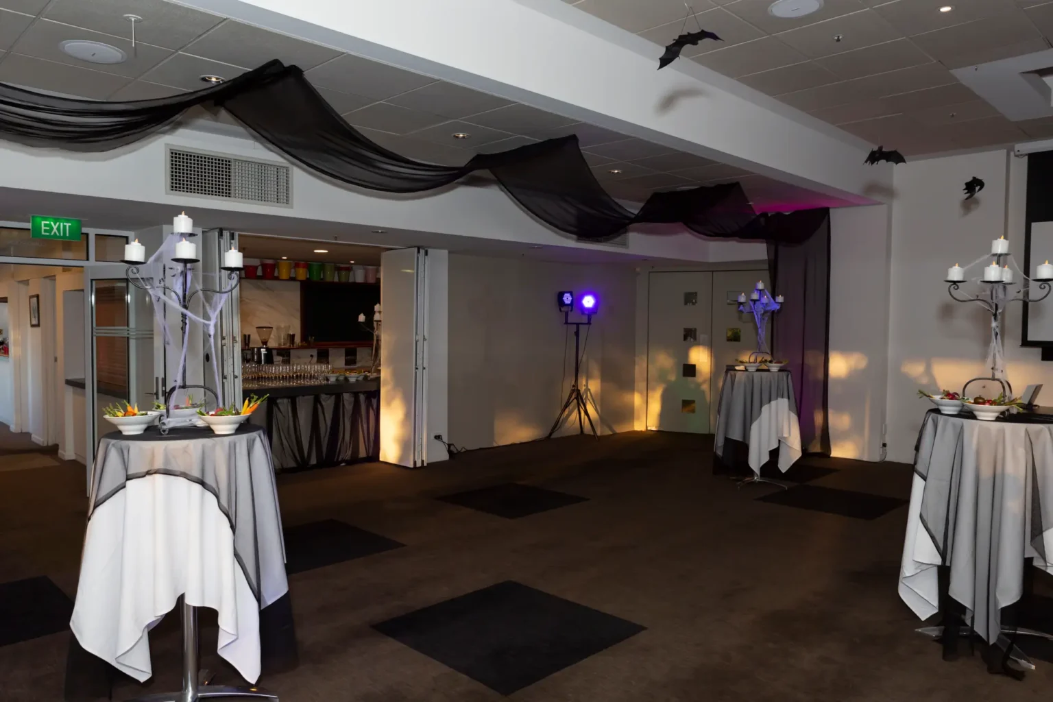 A spacious function room at the Kooyong Tennis Club Halloween Gala is styled with high-top cocktail tables draped in white linens and black mesh overlays, each topped with candelabra centerpieces draped in spiderwebs and bowls of fresh vegetable crudités, beneath black ceiling drapes and bat decorations, while purple, blue, and amber lighting create a moody, sophisticated, and immersive haunted mansion atmosphere in this luxury Melbourne end-of-year ball.