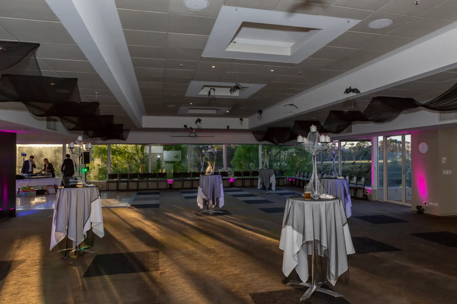A professionally styled ballroom at the Kooyong Tennis Club Halloween Gala shows high cocktail tables with candelabra centrepieces draped in spiderwebs, beneath black ceiling drapes and bat decorations, while magenta lighting washes the walls and golden hour light streams through large windows, with staff preparing in the background, capturing the refined yet spooky atmosphere of this luxury haunted mansion themed event in Melbourne.