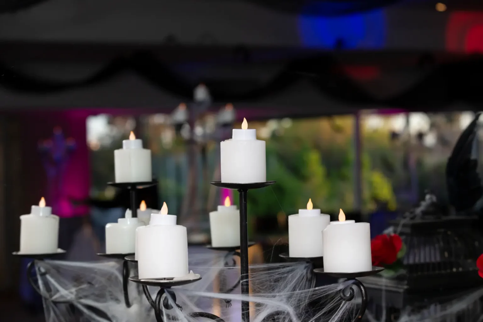 A haunted mansion-inspired centerpiece at the Kooyong Tennis Club Halloween Gala features a black wrought-iron candelabra with glowing LED pillar candles, draped in thick spiderwebs with small black spiders, alongside a dark ornate birdcage accented with deep red roses, while soft magenta and blue lighting and sheer black ceiling drapes create an elegant, eerie atmosphere that captures the gothic romance and luxury of this Melbourne end-of-year ball.