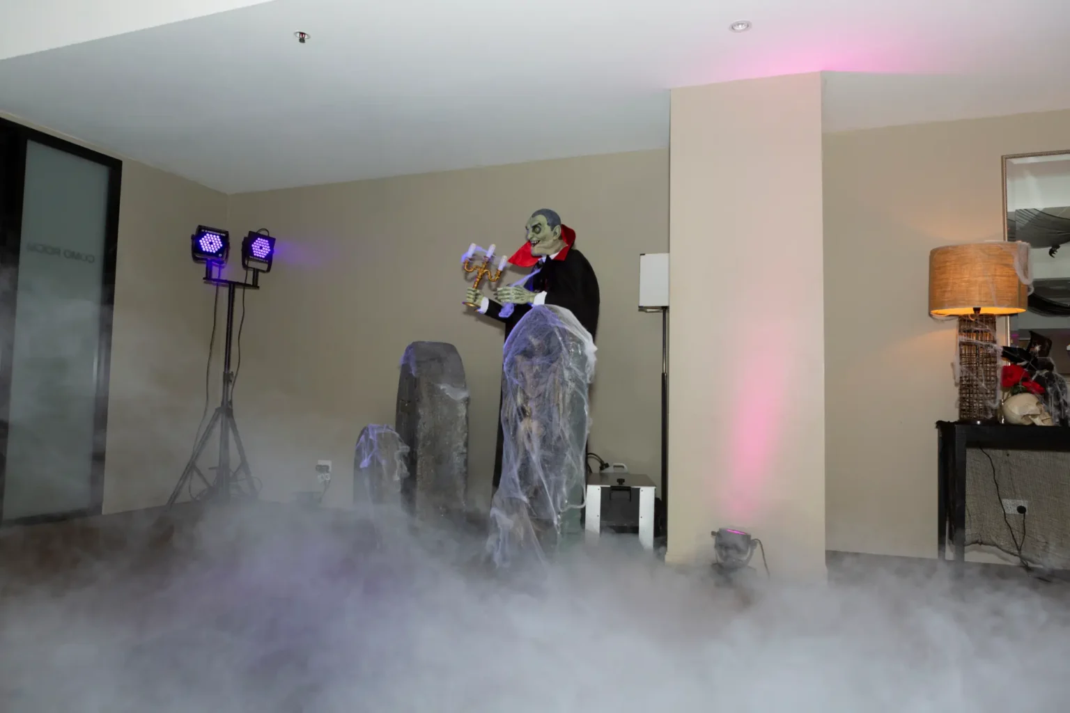 An indoor graveyard scene at the Kooyong Tennis Club Halloween Gala features a life-sized vampire animatronic in a tuxedo and red-lined cape holding a glowing candelabra, surrounded by cobweb-covered tombstones and atmospheric fog drifting across the floor, while purple, blue, and pink lighting enhances the eerie mood, creating an immersive haunted mansion welcome inside this luxury end-of-year ball in Melbourne.