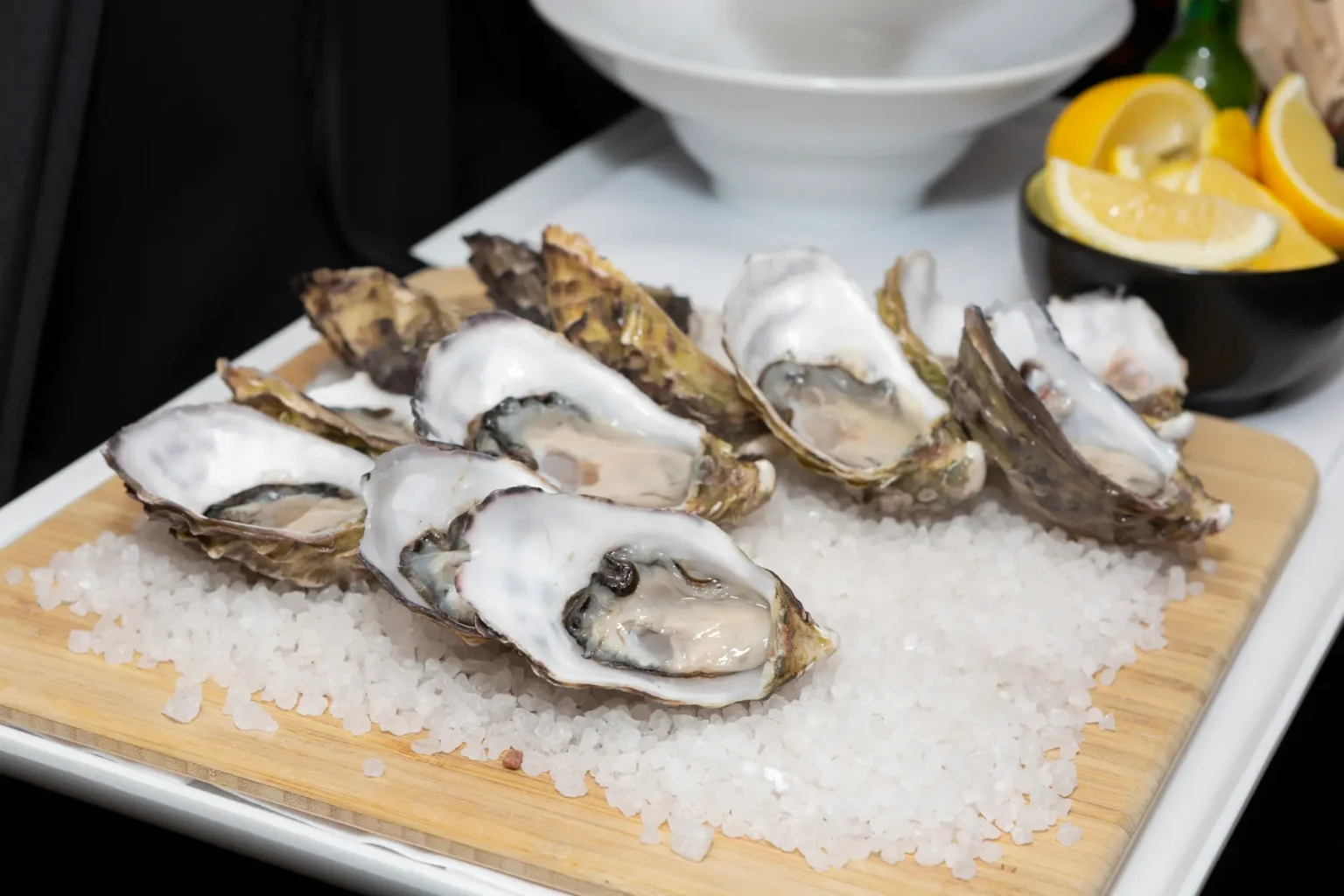 A refined oyster station at the Kooyong Tennis Club Halloween Gala features freshly shucked oysters on the half shell nestled in coarse sea salt on a wooden board and white platter, with lemon wedges in a black bowl and a glimpse of a tuxedoed server nearby, capturing the elegant, high-end catering and luxury atmosphere of this Melbourne end-of-year ball.