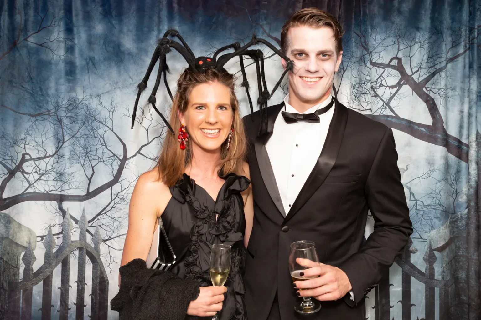 A couple poses at the Kooyong Tennis Club Halloween Gala, Royal South Yarra, blending glamour with spooky-chic flair. The Spider Queen wears a black ruffled dress with a large spider headpiece and red earrings, while the Ghoul dons a black tuxedo with pale "undead" makeup. They stand in front of a misty graveyard backdrop under crisp professional flash lighting.