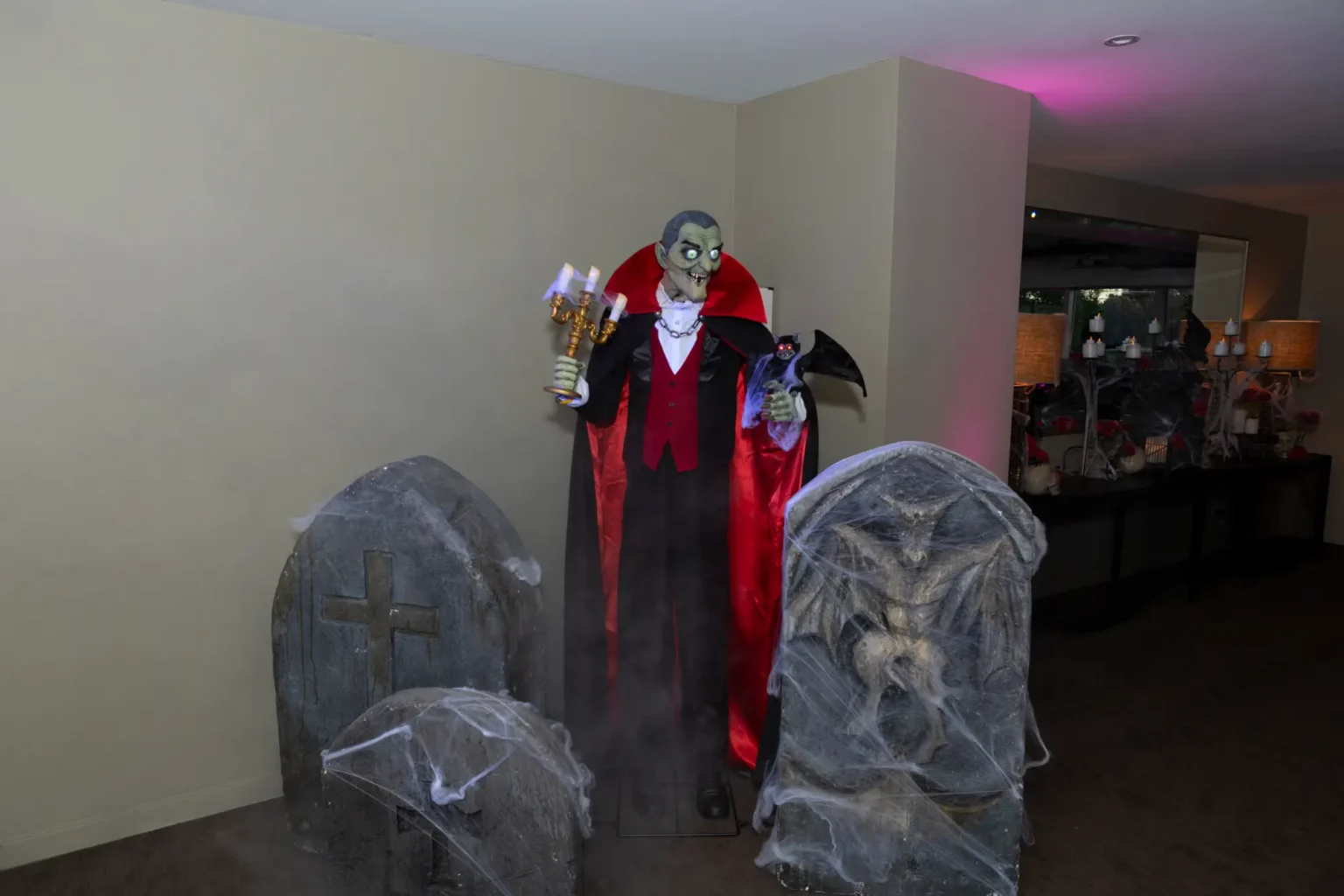 A theatrical graveyard photo-op at the Kooyong Tennis Club Halloween Gala features a life-sized vampire animatronic in a black tuxedo and red-lined cape holding a glowing candelabra and bat, flanked by three spiderweb-draped tombstones, with low-lying mist swirling around the base and soft pink accent lighting reflecting in a nearby mirror, creating an immersive, spooky, and luxurious haunted experience for guests at this Melbourne end-of-year ball.